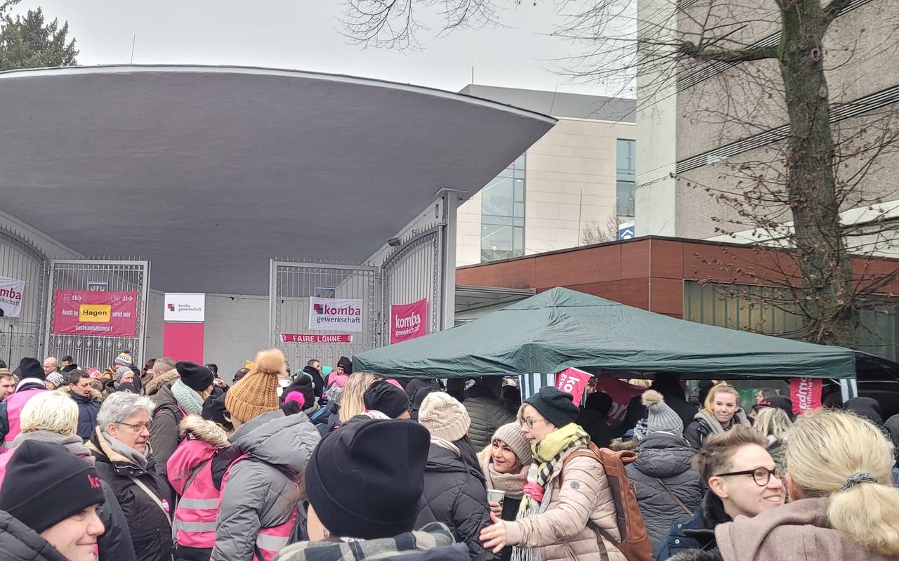 Auf dem Bild sieht man die Konzertmuschel am Volkspark Hagen, sowie den Platz davor. An der Konzertmuschel hängen mehrere pinke Plakate der komba Gewerkschaft mit Sprüchen wie "Auch in Hagen sind wir systemrelevant" oder "Faire Löhne". Auf der Bühne steht noch keiner, dafür ist vor der Konzertmuschel eine Menschenmasse versammelt sowie ein Zelt mit einem Stand aufgebaut. Manche der versammelten Menschen tragen pinke Vesten der komba, andere sind in Alltagskleidung bekleidet.