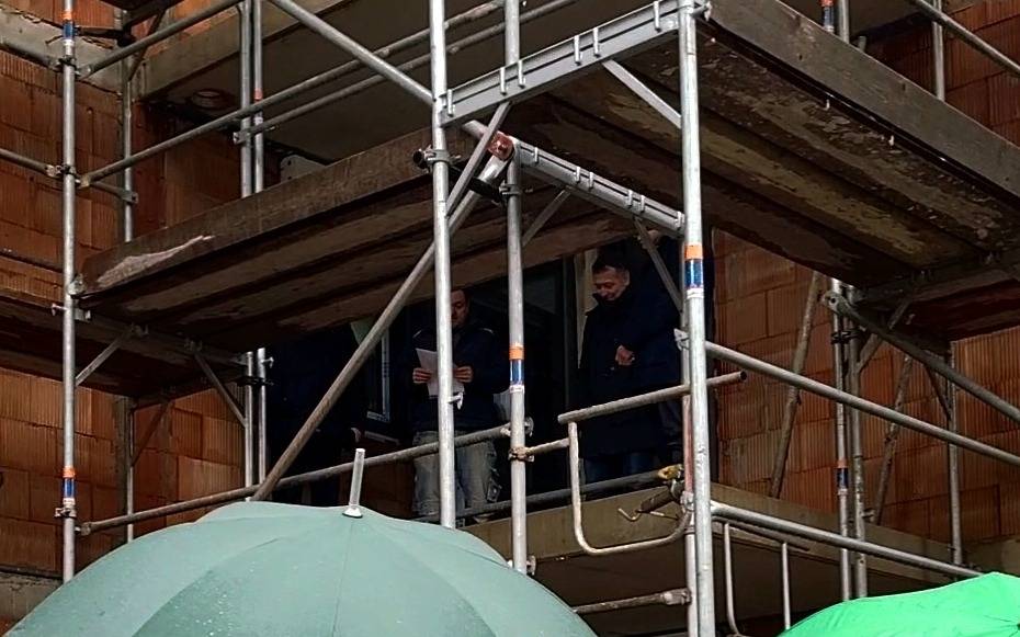 Auf dem Bild zu sehen sind Menschen mit Regenschirmen vor einem Rohbau eines Hauses. An der Baustelle befinden sich Gerüste und Holzplanken. Auf dem Gerüst stehen ebenfalls Menschen. Einer hält Papier in der Hand von dem er den Richtspruch für das Richtfest vorliet.
