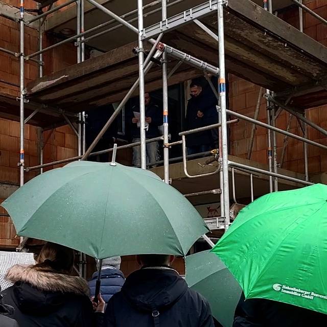 Auf dem Bild zu sehen sind Menschen mit Regenschirmen vor einem Rohbau eines Hauses. An der Baustelle befinden sich Gerüste und Holzplanken. Auf dem Gerüst stehen ebenfalls Menschen. Einer hält Papier in der Hand von dem er den Richtspruch für das Richtfest vorliet.