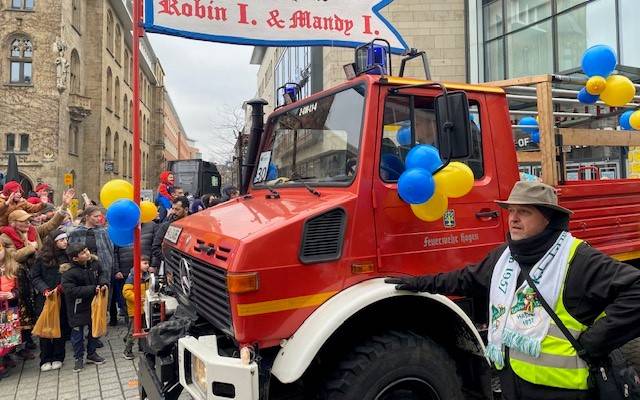 Ein festlich geschmücktes Feuerwehrauto fährt beim Rosenmontagsumzug durch die Innenstadt. Auf dem Auto ist das diesjährige Prinzenpaar. Am Feuerwehrauto ist ein Banner mit den Namen des Paares.