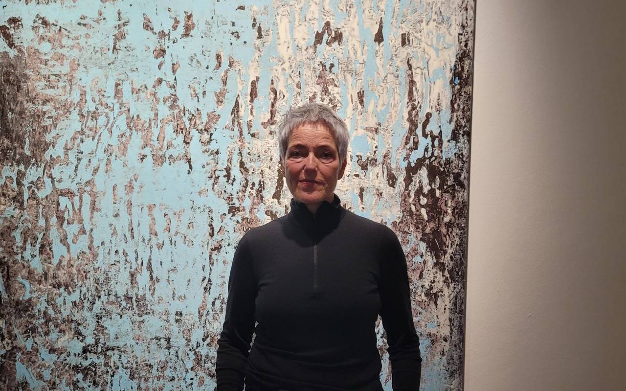 Auf dem Bild ist eine Frau in Alltagskleidung zu sehen. Die Künstlerin, Andrea Behn, steht vor einem Kunstwerk von ihr auf Papier. Das Kunstwerk besteht aus abstrakten blau-grauen Farbschichten und ist in einem großen Format, welches ihre Körpergröße überragt.