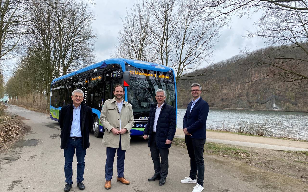 Vier Männer stehen in einem Halbkreis. Hinter ihnen sieht man einen Bus und einen See.