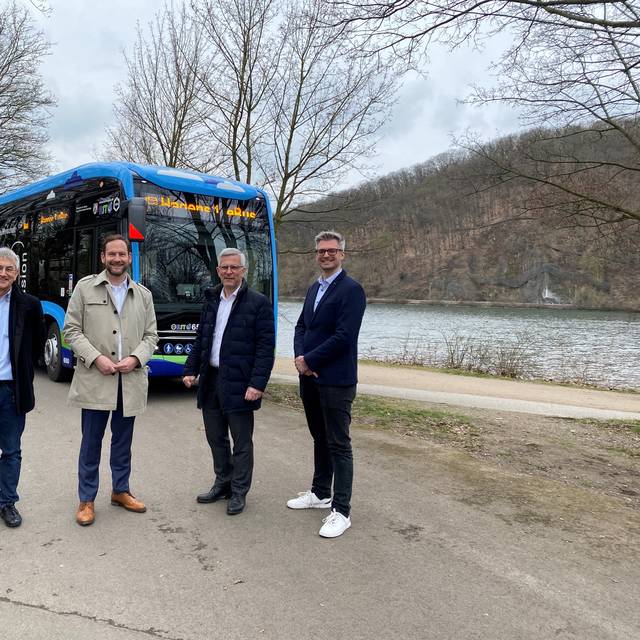Vier Männer stehen in einem Halbkreis. Hinter ihnen sieht man einen Bus und einen See.