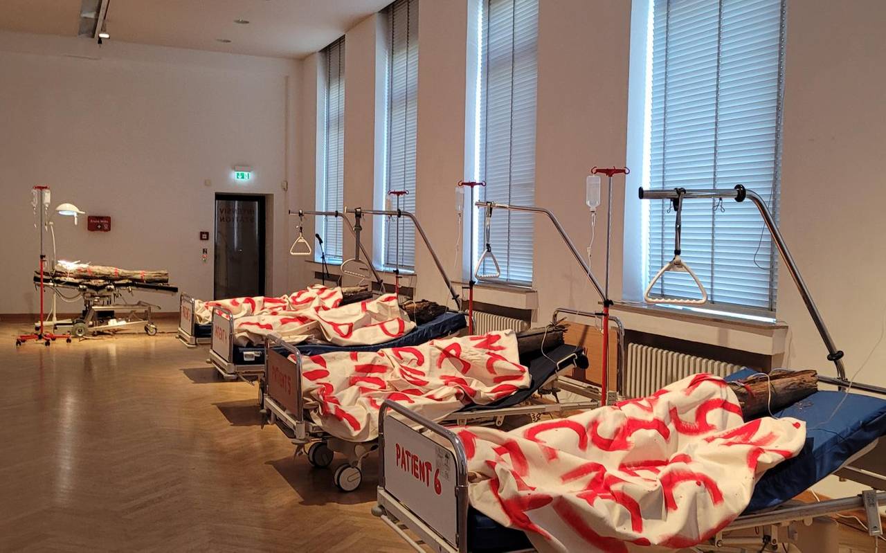Zu sehen sind Baumstämme, die auf Krankenbetten platziert sind und mit Leinen bedeckt sind, auf denen in rot Begriffe wie "Emergency" oder "Help" stehen.