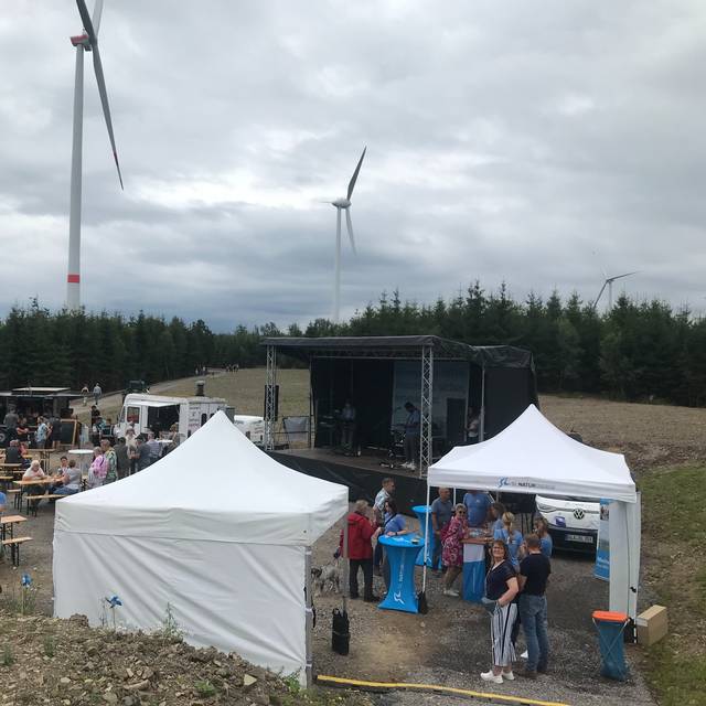 Im Hintergrund stehen mehrere Windräder. Vorne sind Zelte aufgebaut und Bierzeltgarnituren. Darauf sitzen Menschen.