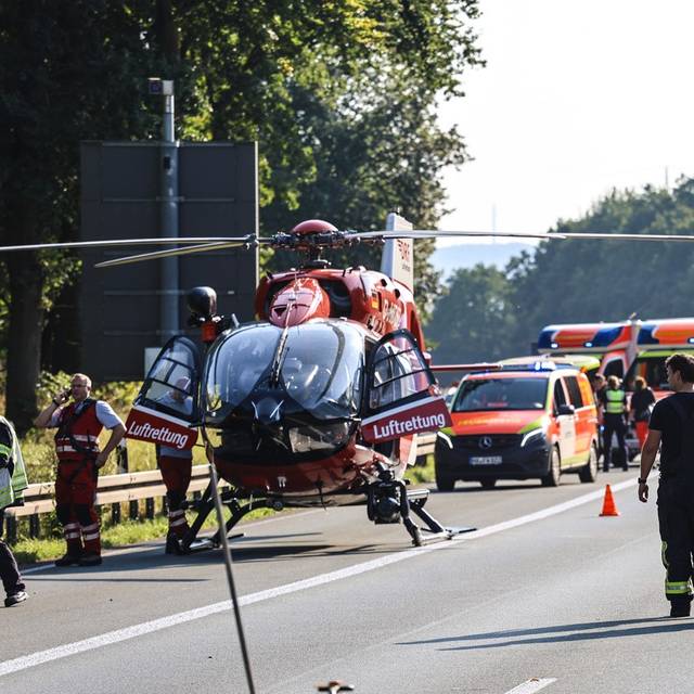 Ein roter Rettungshubschrauber steht auf der Straße. Neben ihm stehen zwei Feuerwehrleute. Im Hintergrund sieht man mehrere Rettungsfahrzeuge.