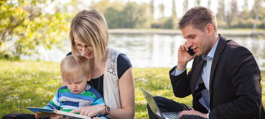 Eine junge Familie picknickt draußen in der Natur, während der Mann Büroarbeiten am Laptop erledigt.