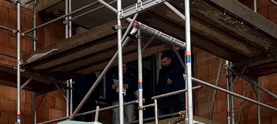 Auf dem Bild zu sehen sind Menschen mit Regenschirmen vor einem Rohbau eines Hauses. An der Baustelle befinden sich Gerüste und Holzplanken. Auf dem Gerüst stehen ebenfalls Menschen. Einer hält Papier in der Hand von dem er den Richtspruch für das Richtfest vorliet.