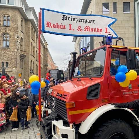 Ein festlich geschmücktes Feuerwehrauto fährt beim Rosenmontagsumzug durch die Innenstadt. Auf dem Auto ist das diesjährige Prinzenpaar. Am Feuerwehrauto ist ein Banner mit den Namen des Paares.