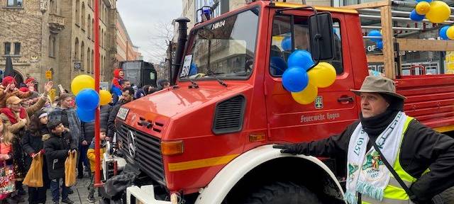 Ein festlich geschmücktes Feuerwehrauto fährt beim Rosenmontagsumzug durch die Innenstadt. Auf dem Auto ist das diesjährige Prinzenpaar. Am Feuerwehrauto ist ein Banner mit den Namen des Paares.