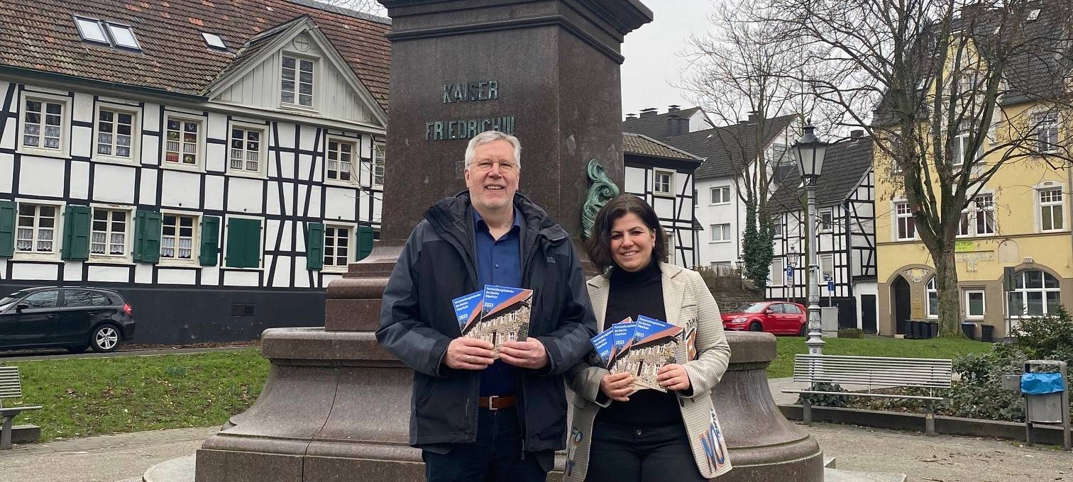 Der Bezirksbürgermeister Michael Dahme und die Geschäftsstellenmitarbeiterin Züleyha Kandemir stehen in Eilpe/Dahl vor dem Denkmal. In den Händen halten sie den frisch gedruckten Veranstaltungskalender.