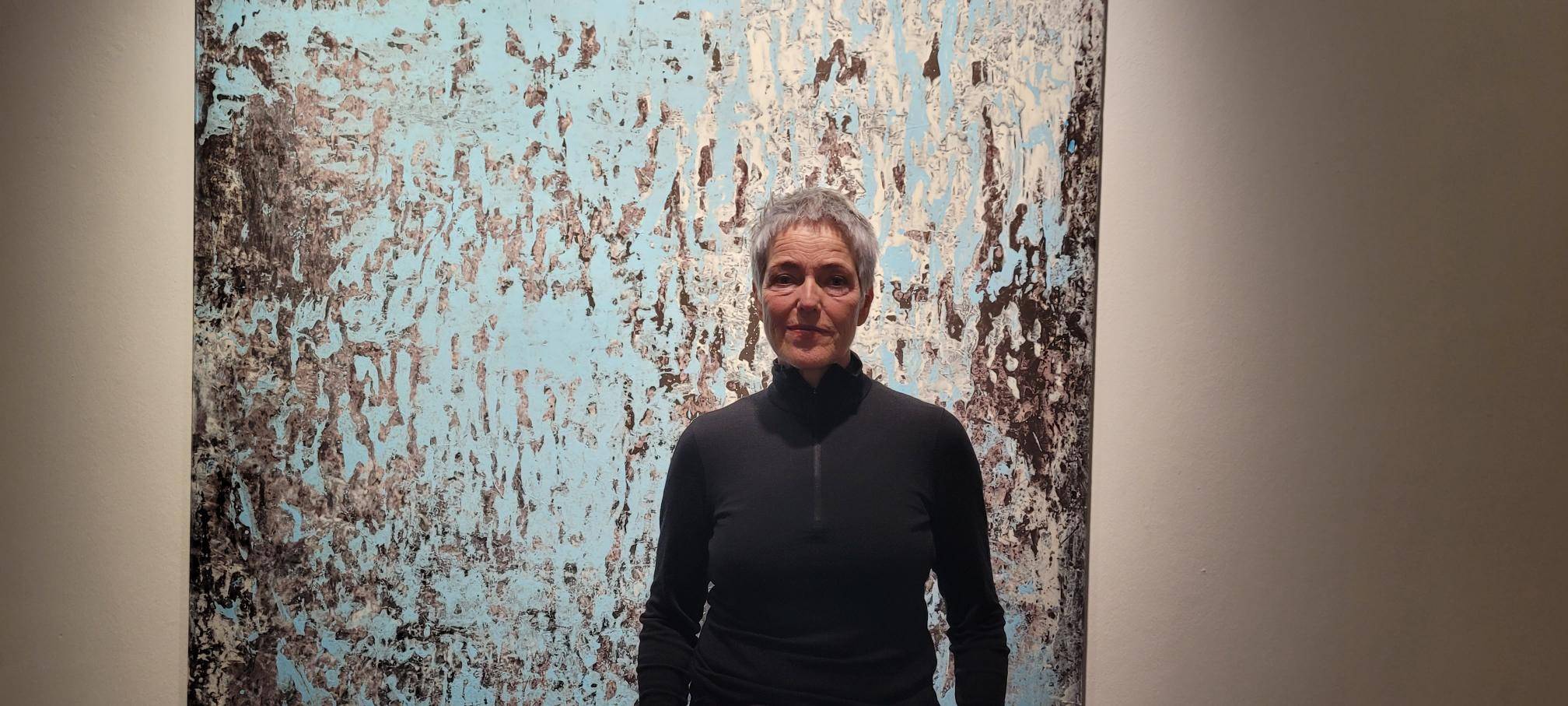 Auf dem Bild ist eine Frau in Alltagskleidung zu sehen. Die Künstlerin, Andrea Behn, steht vor einem Kunstwerk von ihr auf Papier. Das Kunstwerk besteht aus abstrakten blau-grauen Farbschichten und ist in einem großen Format, welches ihre Körpergröße überragt.