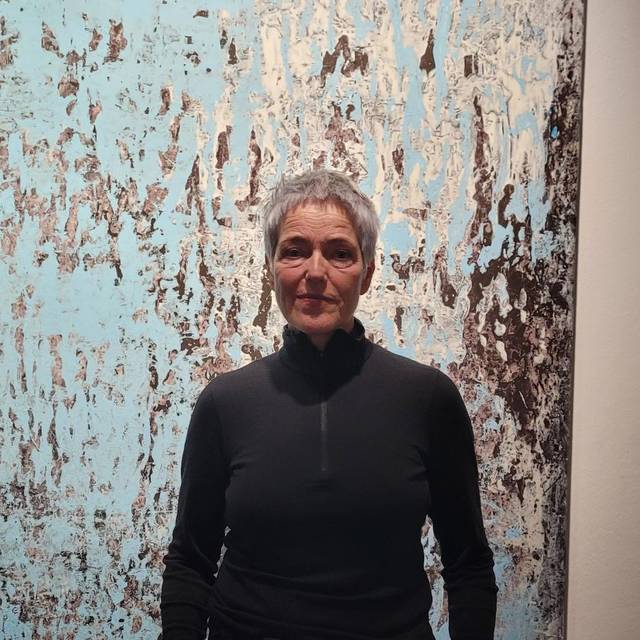 Auf dem Bild ist eine Frau in Alltagskleidung zu sehen. Die Künstlerin, Andrea Behn, steht vor einem Kunstwerk von ihr auf Papier. Das Kunstwerk besteht aus abstrakten blau-grauen Farbschichten und ist in einem großen Format, welches ihre Körpergröße überragt.