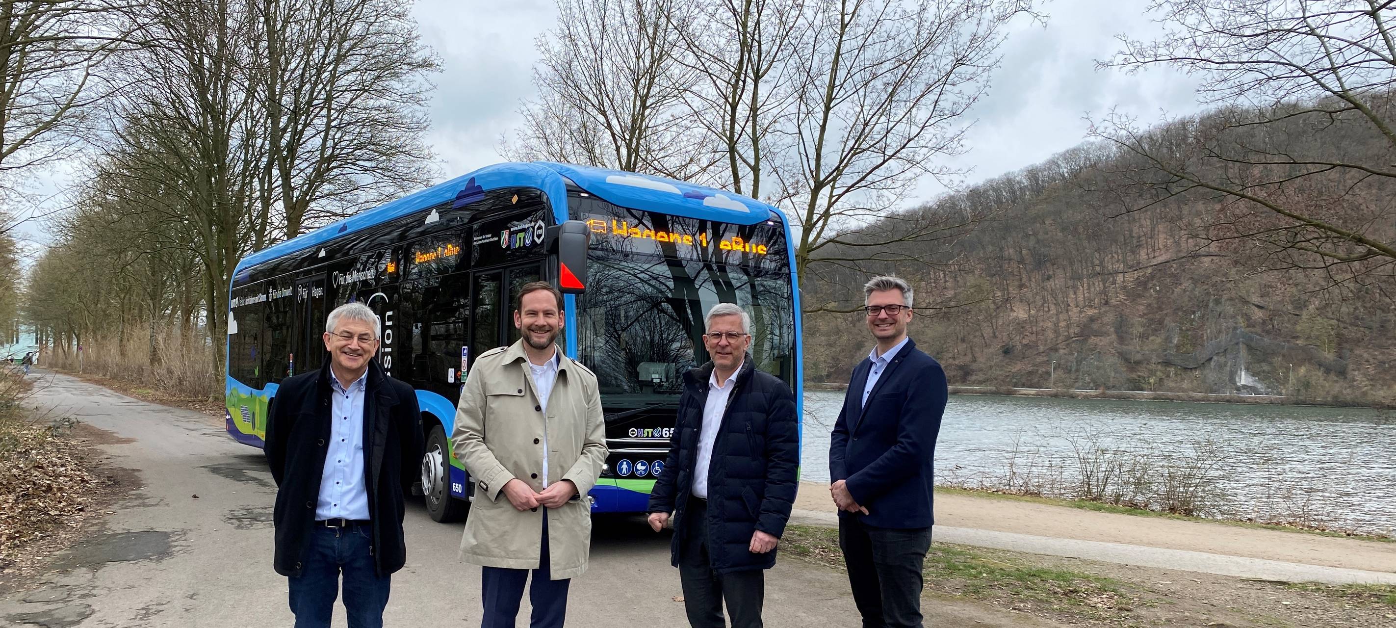 Vier Männer stehen in einem Halbkreis. Hinter ihnen sieht man einen Bus und einen See.