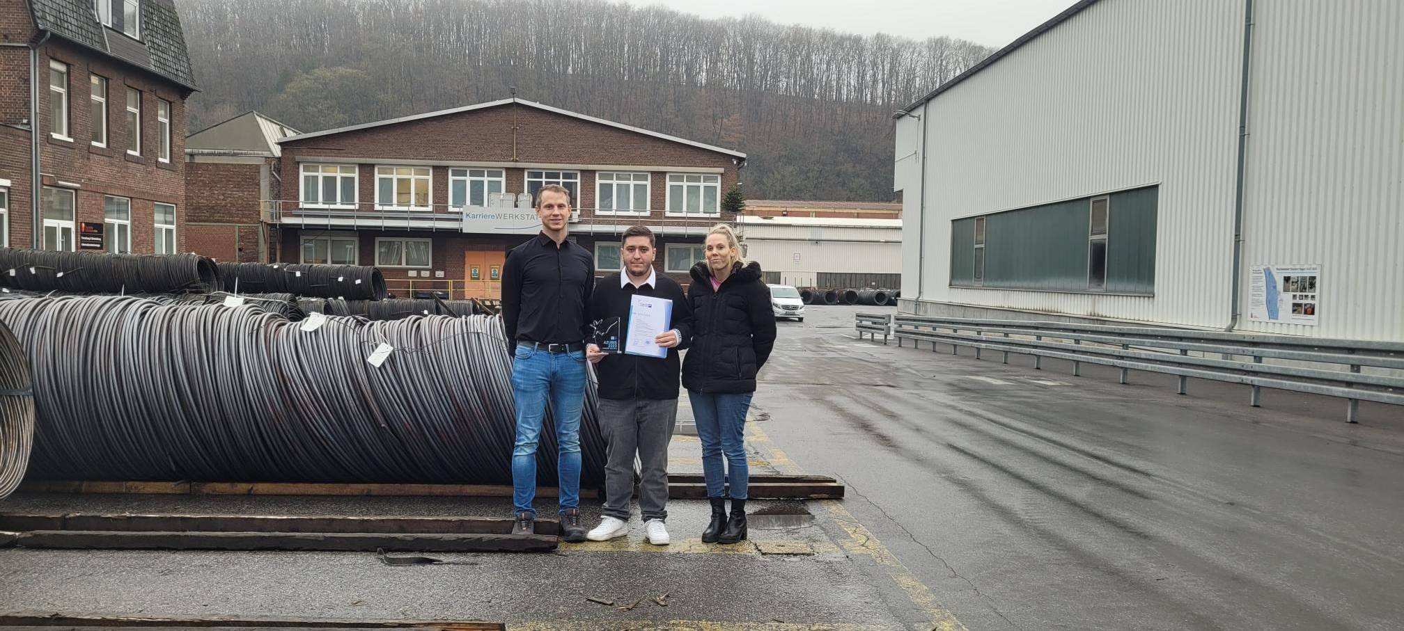 Zu sehen sind 3 Leute vor Stahldrähten bei den Deutschen Edelstahlwerken. Darunter der Ausbildungsleiter Kristian Baum, Vertreterin des Ausbildungsrates und der Azubi Bora Turan, der seinen Preis hält.