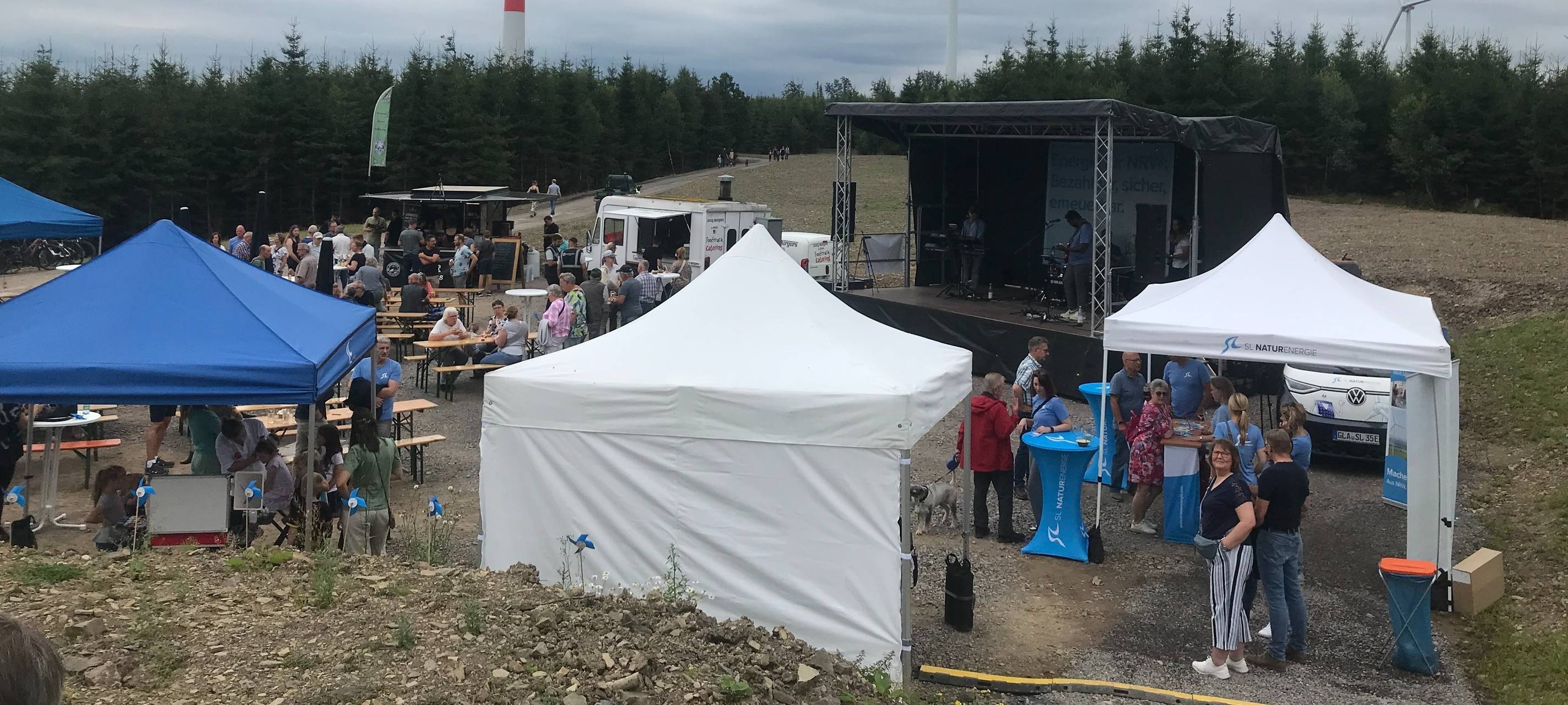 Im Hintergrund stehen mehrere Windräder. Vorne sind Zelte aufgebaut und Bierzeltgarnituren. Darauf sitzen Menschen.