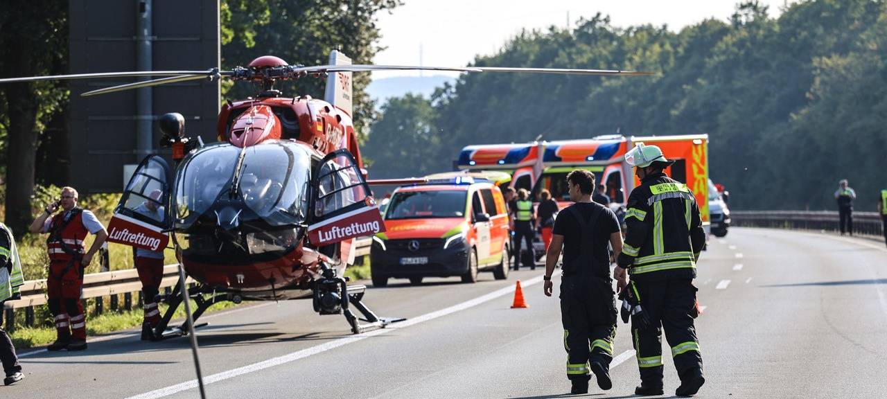 Ein roter Rettungshubschrauber steht auf der Straße. Neben ihm stehen zwei Feuerwehrleute. Im Hintergrund sieht man mehrere Rettungsfahrzeuge.