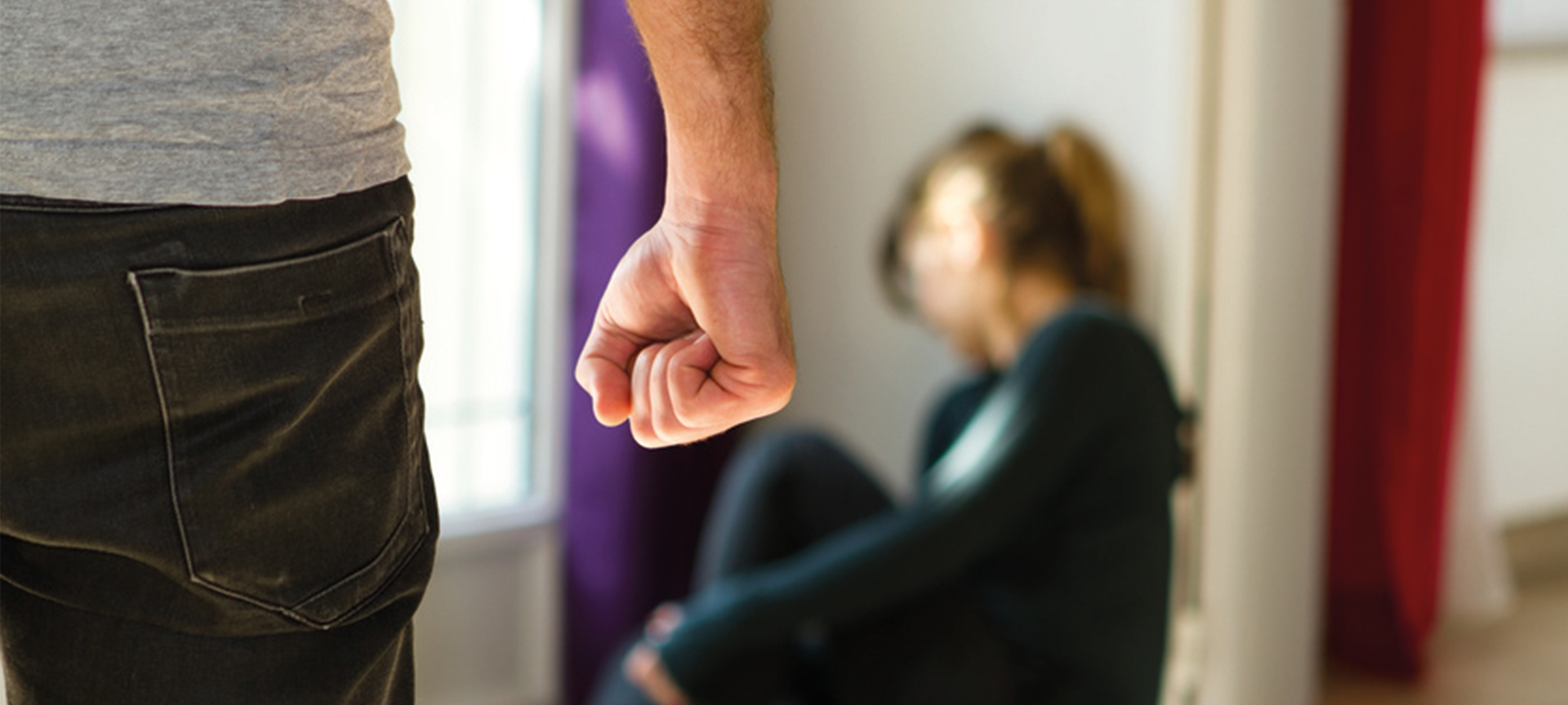 Frau sitzt verängstigt in einer Ecke. Ein Mann hat die Faust geballt.